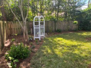 Backyard with a white arbor, wooden fence, grass, and trees.