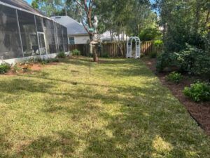Backyard with green grass, screened porch, and garden beds along the edges. A white arched trellis stands near a wooden fence.