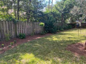 Backyard with a wooden fence, green grass, mulch border, and trees in the background.