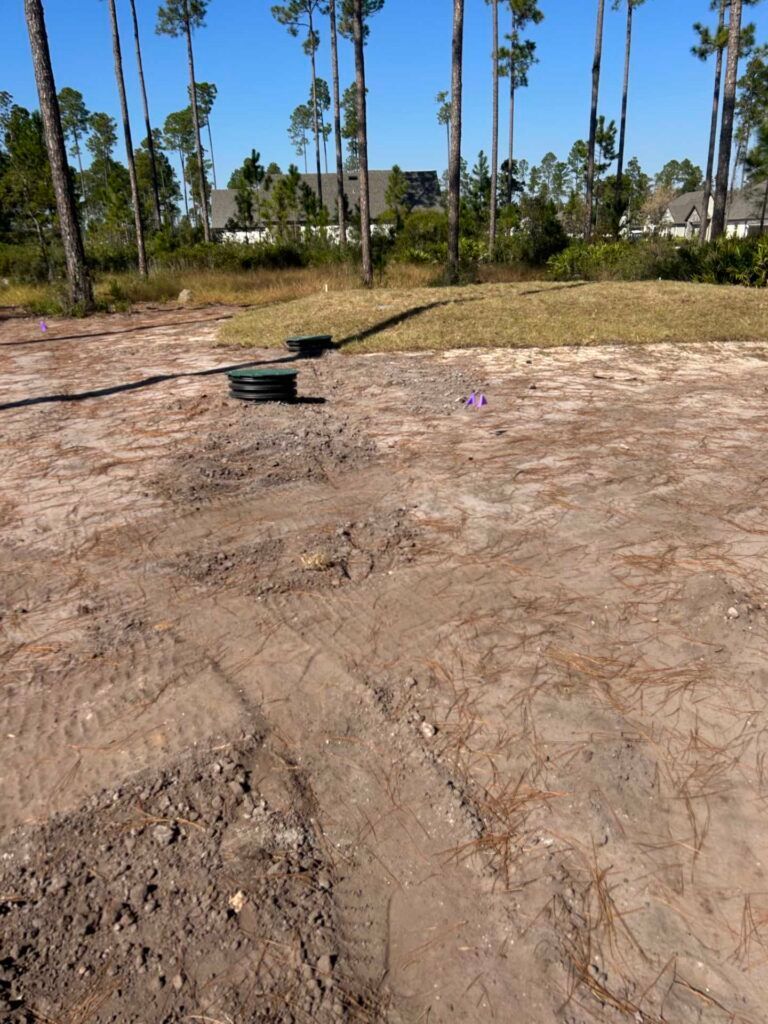Dirt lot with construction markers; trees and houses in the background under a blue sky.