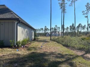 Side view of a house next to a grassy area with a dirt path. In the distance are more houses and tall trees.