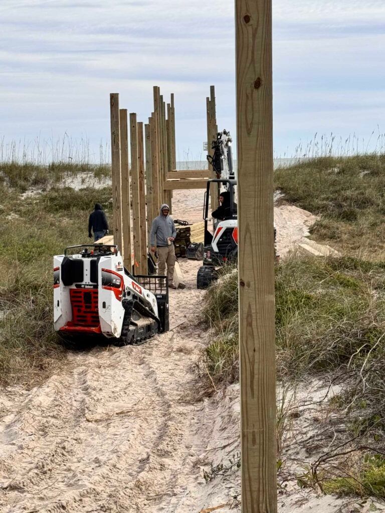 Construction of a wooden structure on a sandy path near the ocean. Two compact construction vehicles, workers present.