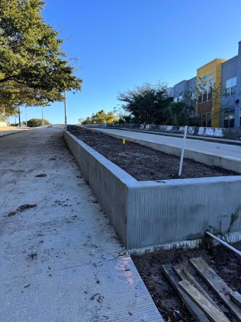 Concrete planters filled with dirt on a sloped gravel path, in front of buildings on a sunny day.