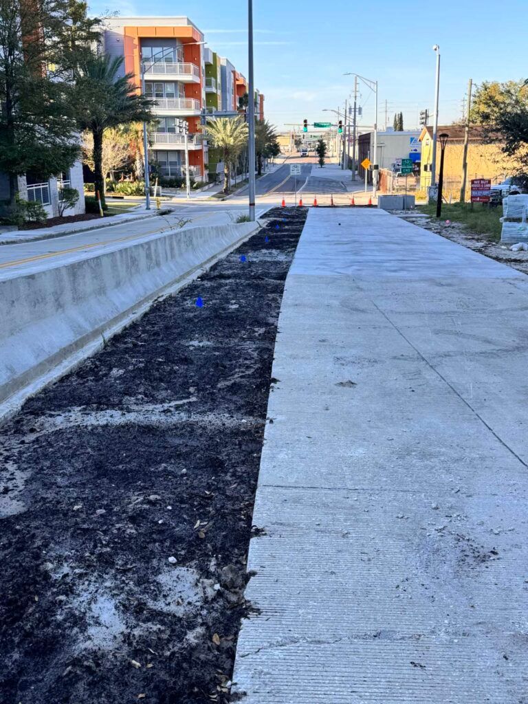 Road construction site; long concrete median with exposed asphalt, road ahead blocked by cones; buildings in background.