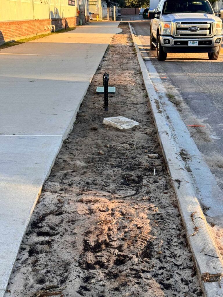 Long dirt strip between sidewalk and street with water pipe and parked truck.