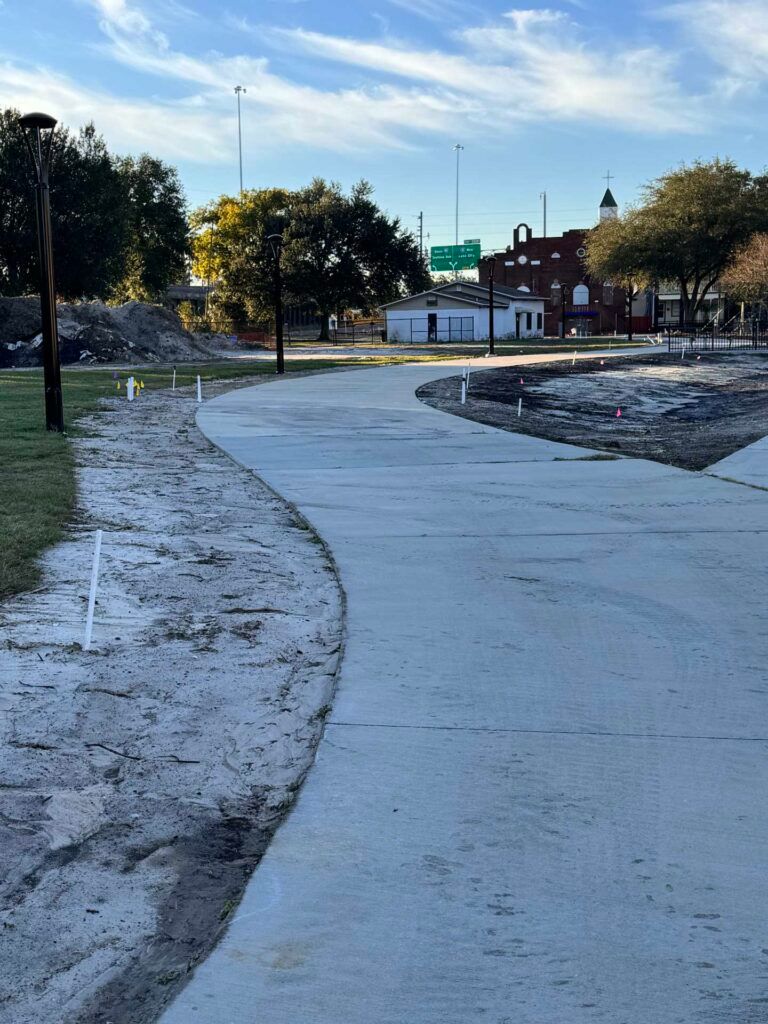 Concrete path curving through a park. Trees, buildings, and construction debris in background under a cloudy sky.