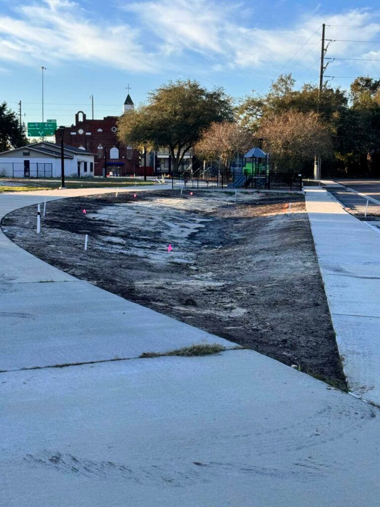 New park area with exposed soil and playground equipment, surrounded by concrete walkways, and trees.