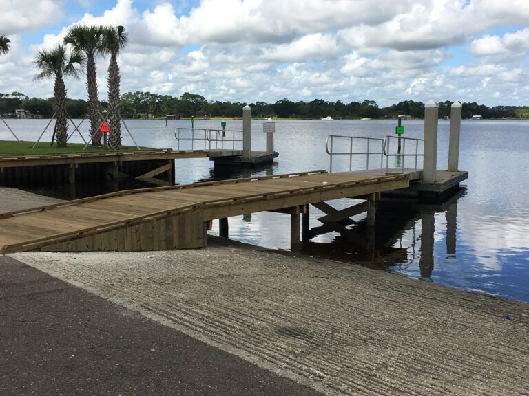 Wooden dock extending into a calm lake under cloudy skies, with white railings and palm trees nearby