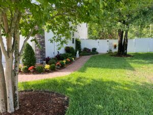 Lush green lawn and brick pathway leading to a white building. Colorful flowers border the path, under shady trees.