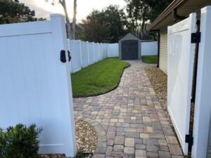 White fence with gates opening to a brick path leading to a shed. Grass and landscaping on the left.