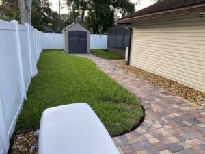 Lawn and brick pathway alongside a white fence and tan house. A shed is in the distance.