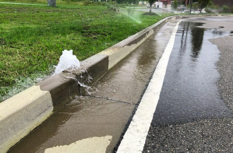 Water gushing from a roadside curb drain onto a wet street beside a grassy embankment