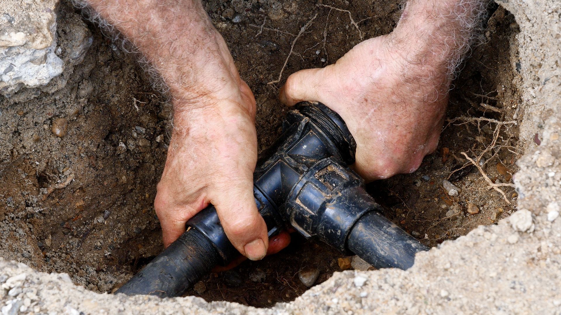 Hands twisting a black plastic pipe fitting in a dirt trench during repair work