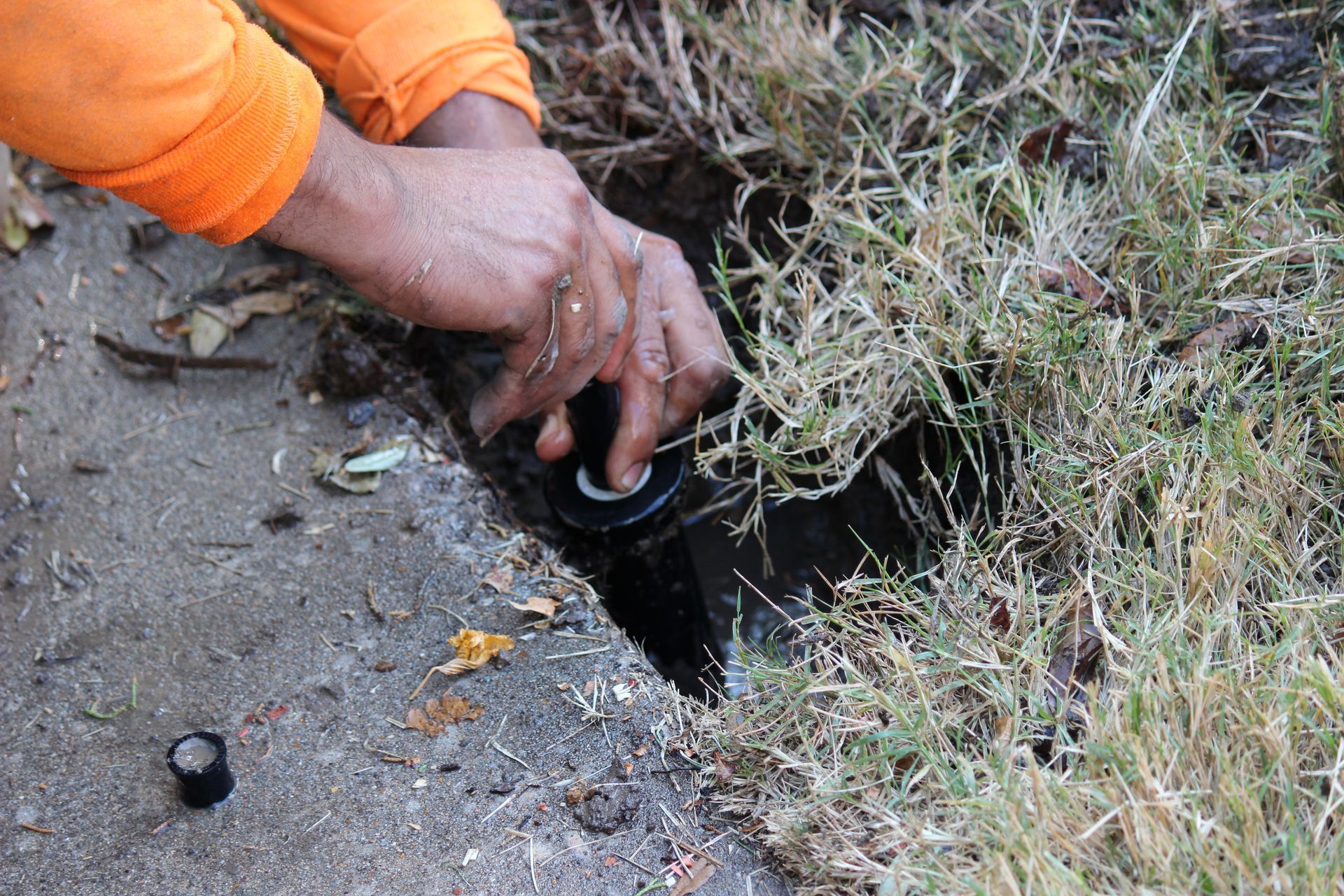 Hands in an orange sleeve working on a black pipe in dirt beside grass.
