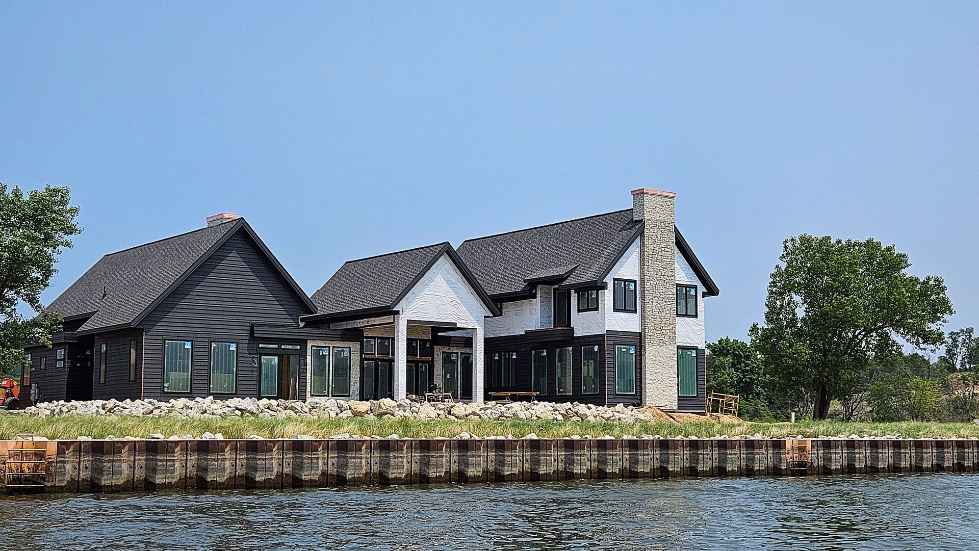 Waterfront house with dark shingled roof and white stone tower beside calm water under a blue sky