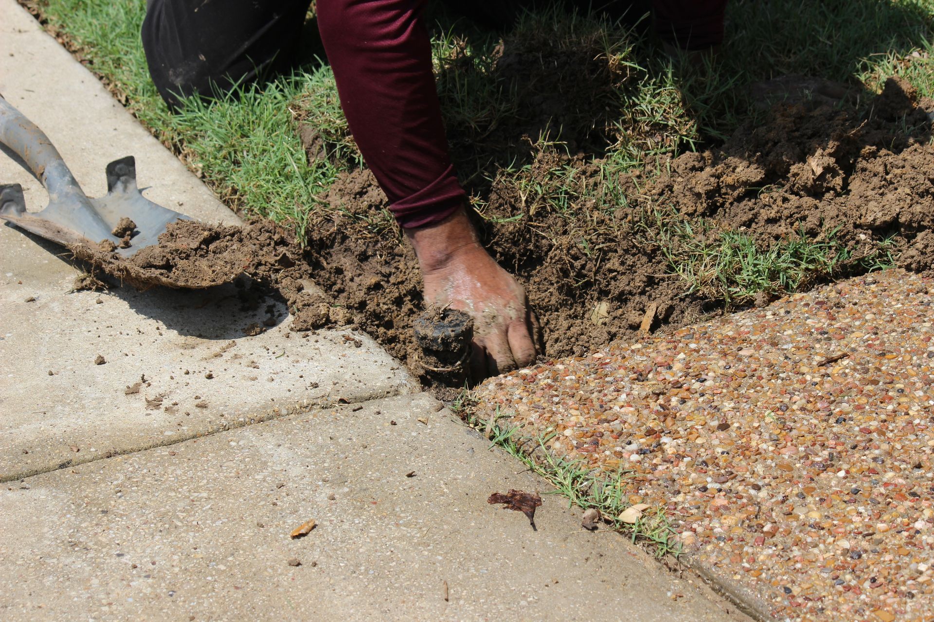Person digging soil beside a sidewalk with a hand tool near a concrete edge and gravel border