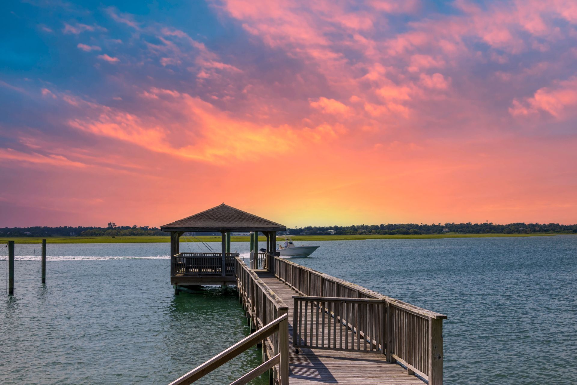 Wooden pier leading to a gazebo over calm water at sunset with a pink-orange sky