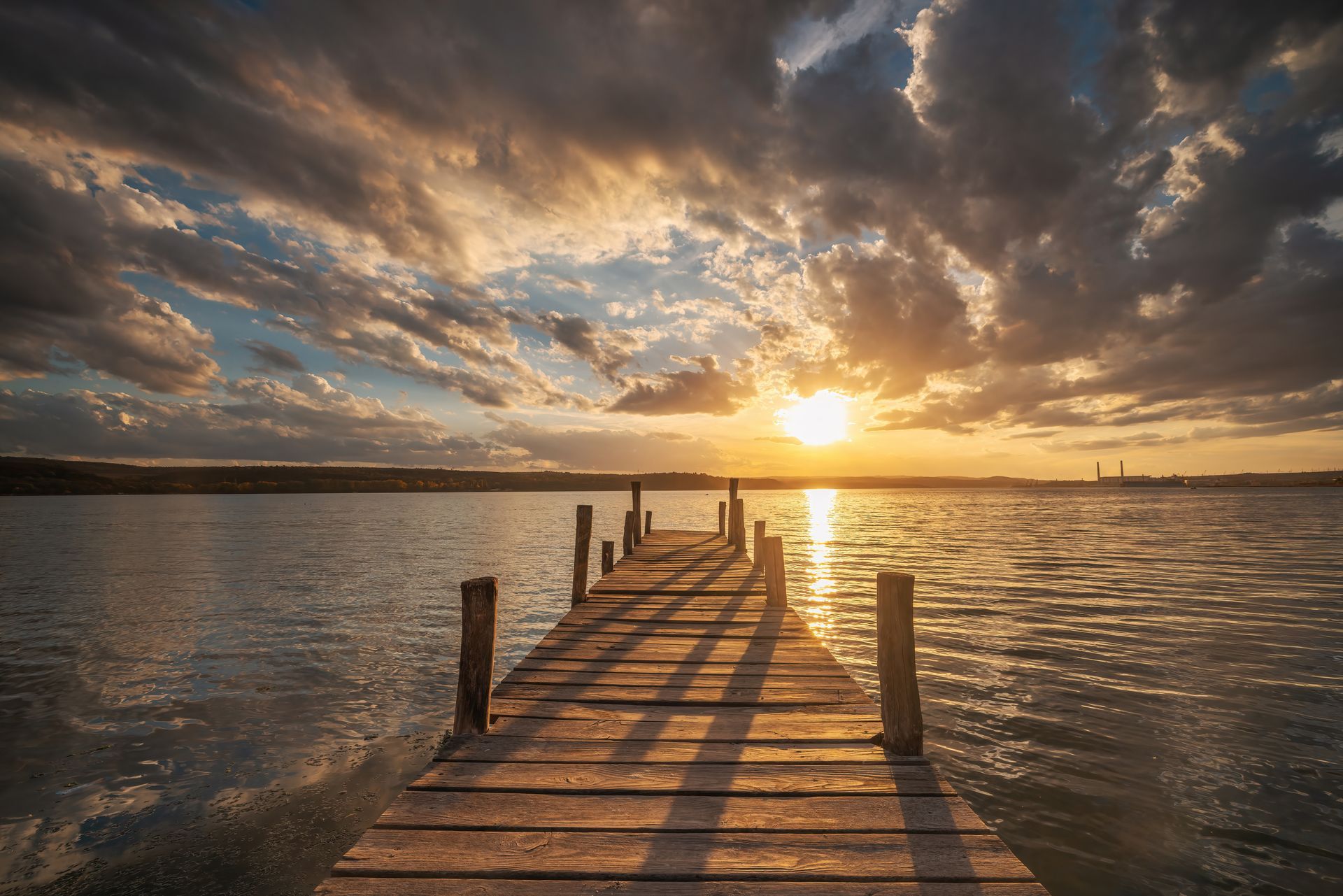 Wooden dock leading into calm water at sunset, with dramatic clouds and golden reflections.