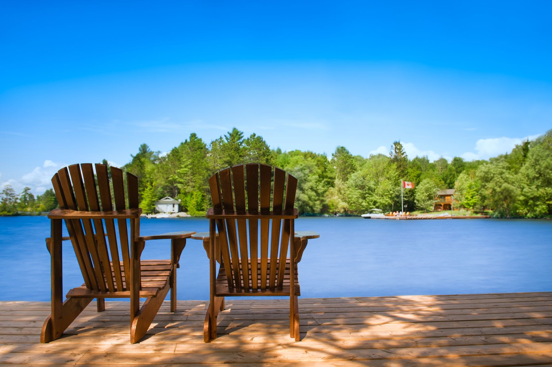 Two wooden chairs on a sandy shore overlooking a calm blue lake and green trees under a clear sky
