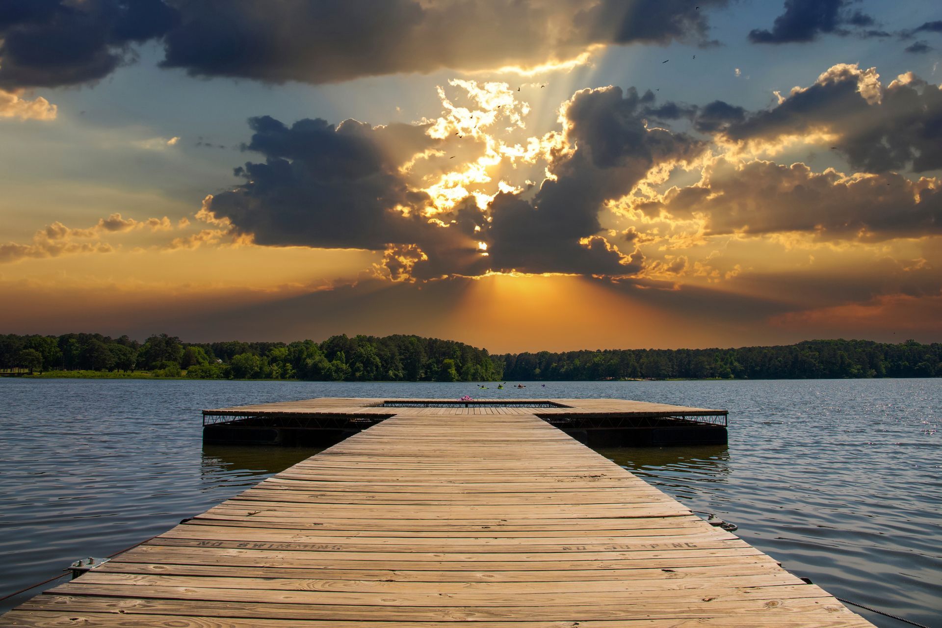 Wooden dock stretching into a calm lake at sunset, with golden clouds and sun rays over distant trees