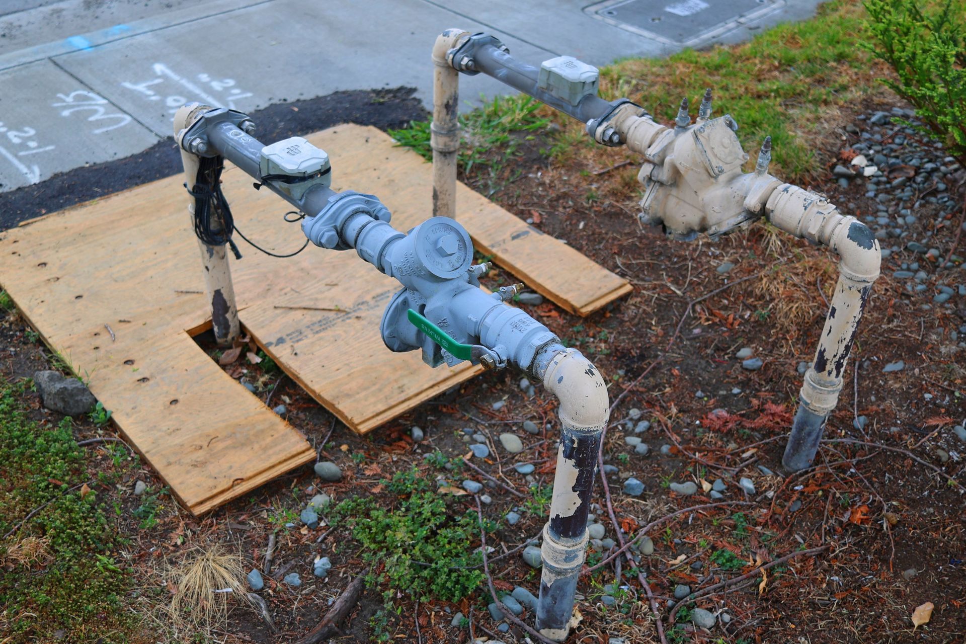 Rusty metal pipe assembly with valves and wooden supports beside pavement and soil