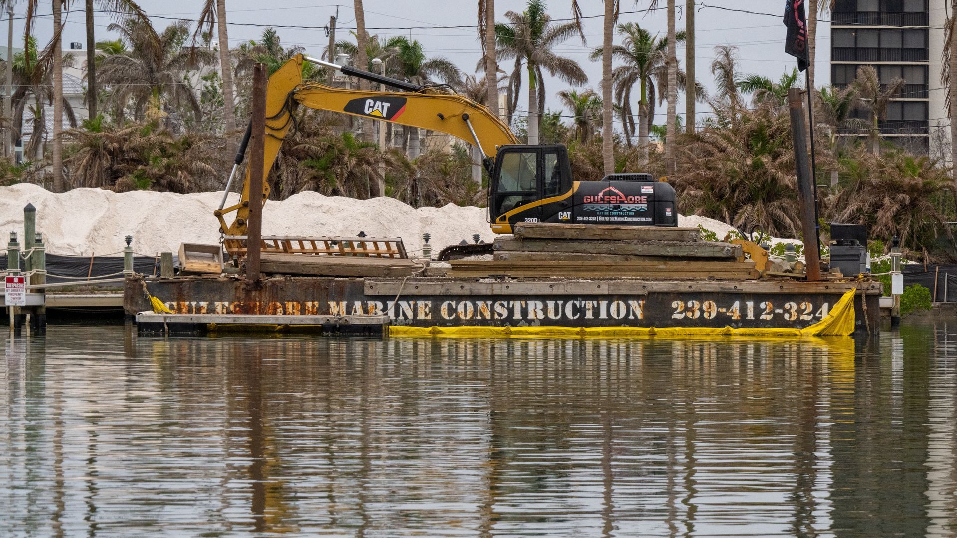 Yellow excavator on a floating barge in calm water, with palm trees and construction materials in the background.