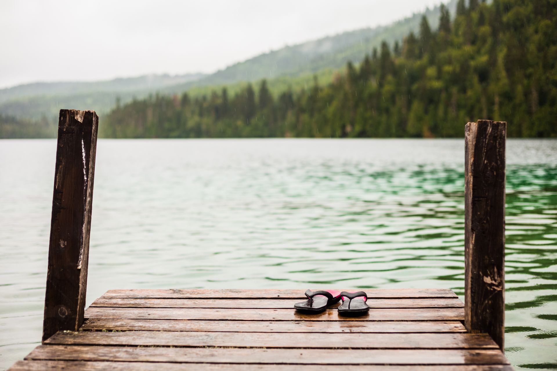 Sandals on a wooden dock by a calm lake with forested mountains in the background