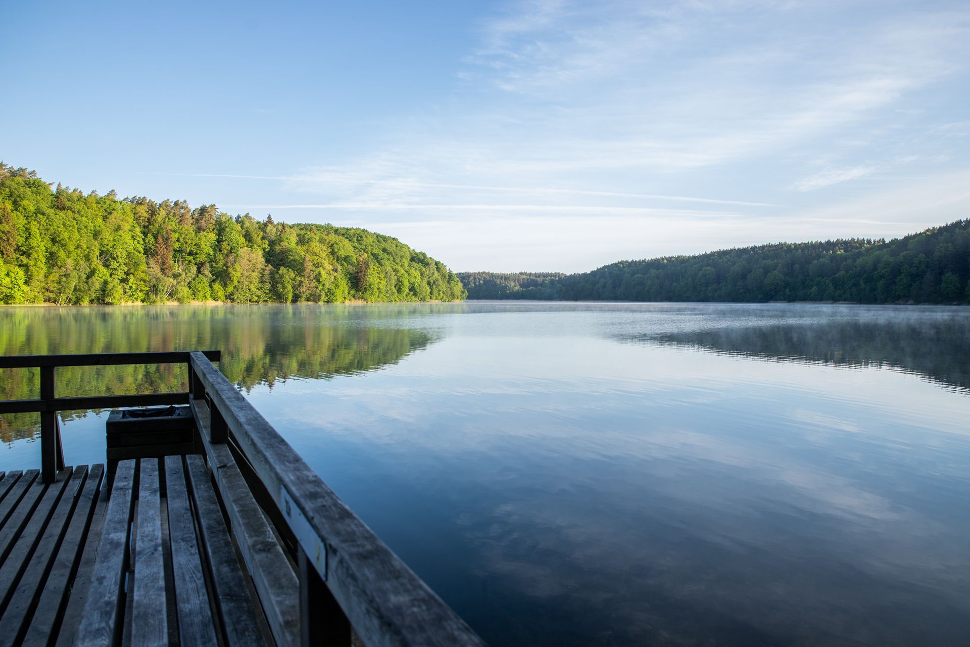 Wooden dock beside a calm lake at sunrise, with green forest and clear reflections.