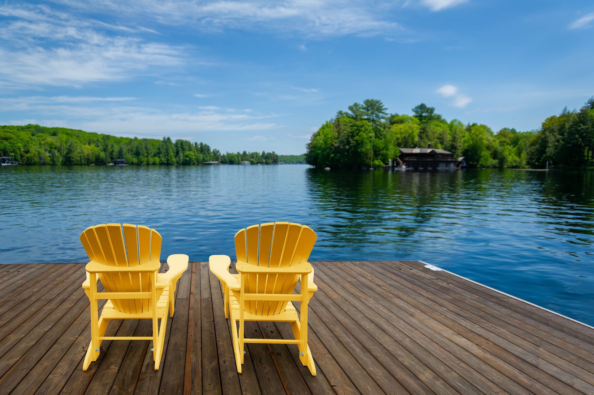 Two yellow Adirondack chairs on a wooden dock overlooking a calm blue lake and green trees