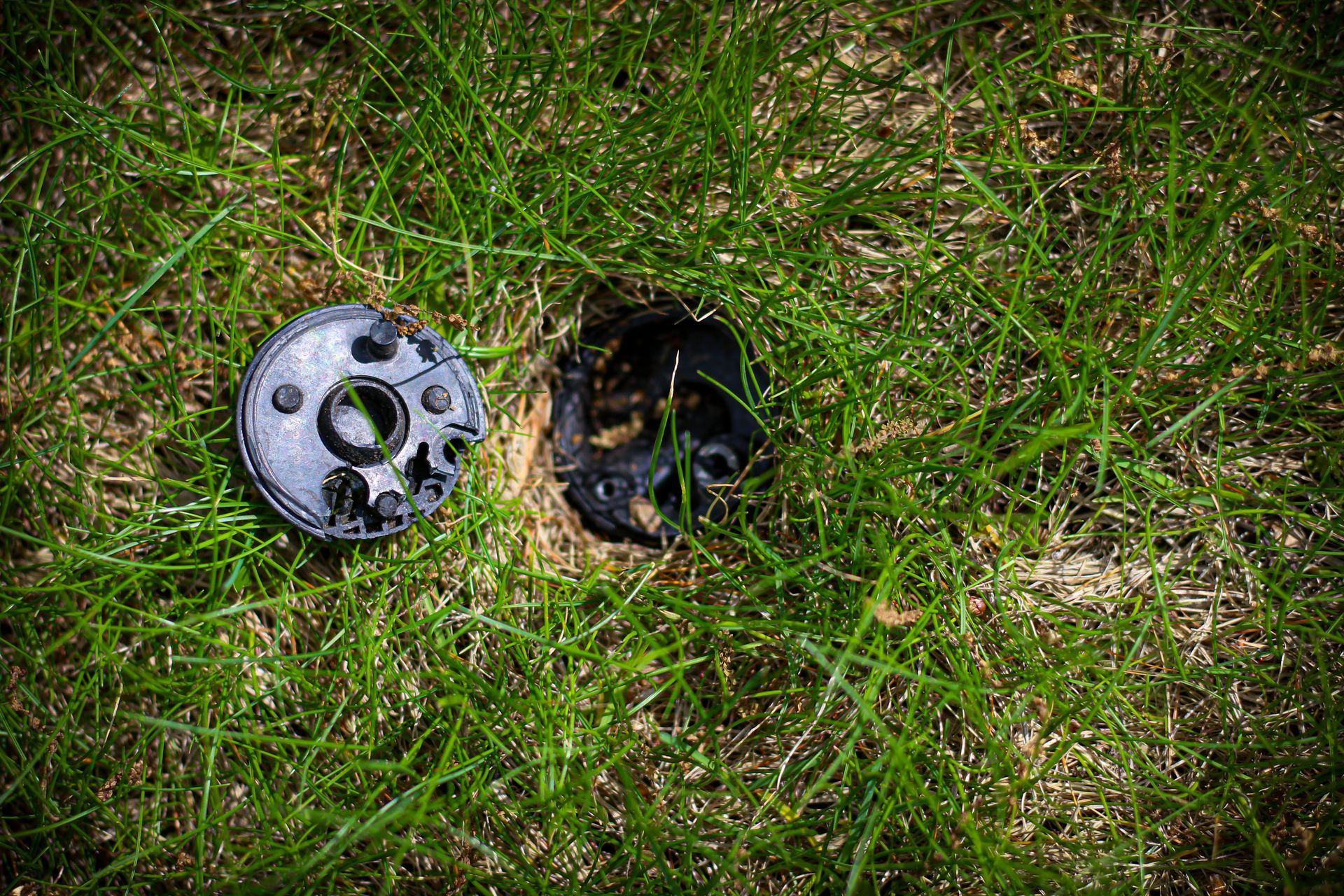 Damaged metal cap and open hole in grass, with burnt soil around the opening