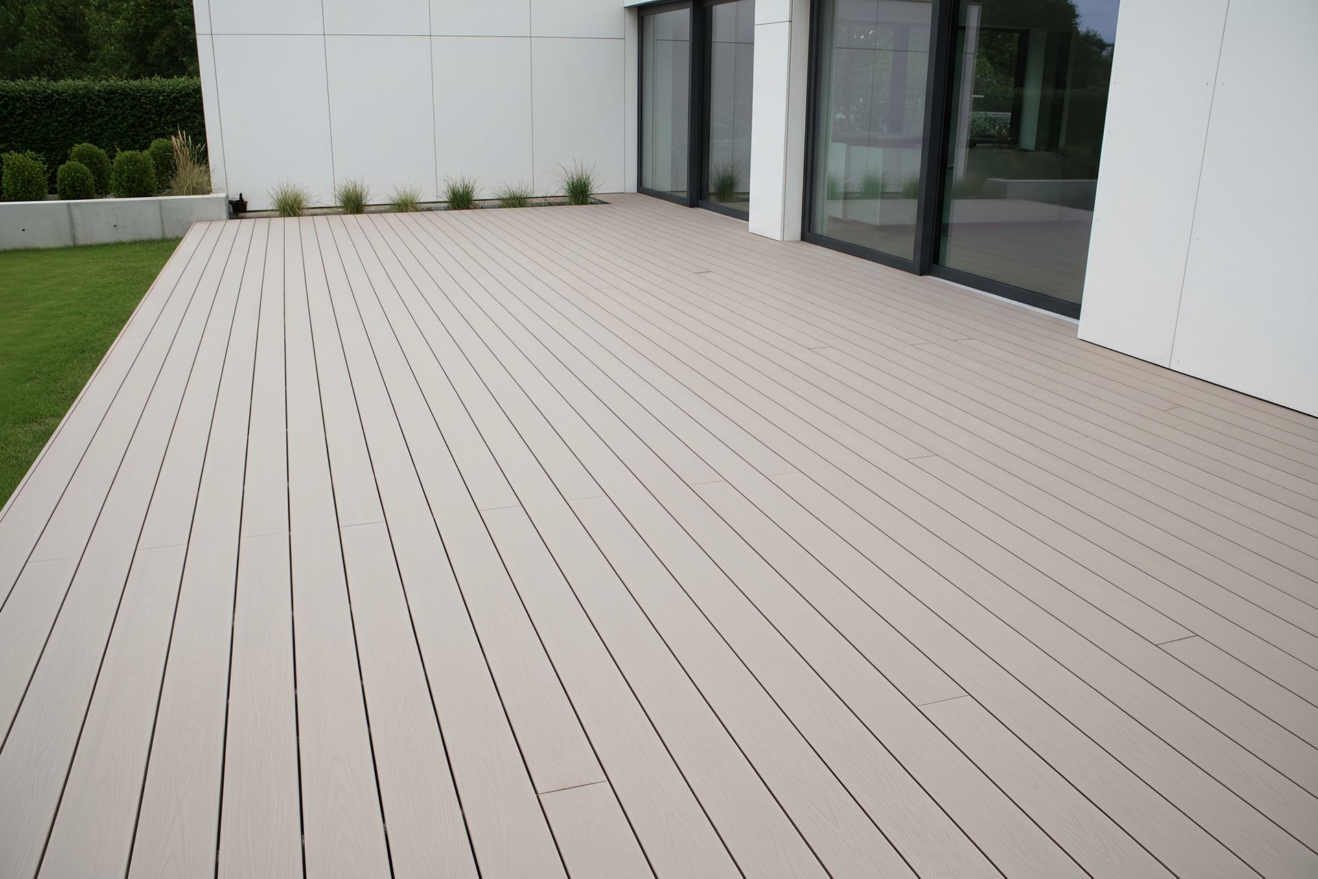 Wide beige outdoor deck beside a modern white building with glass doors and green grass.