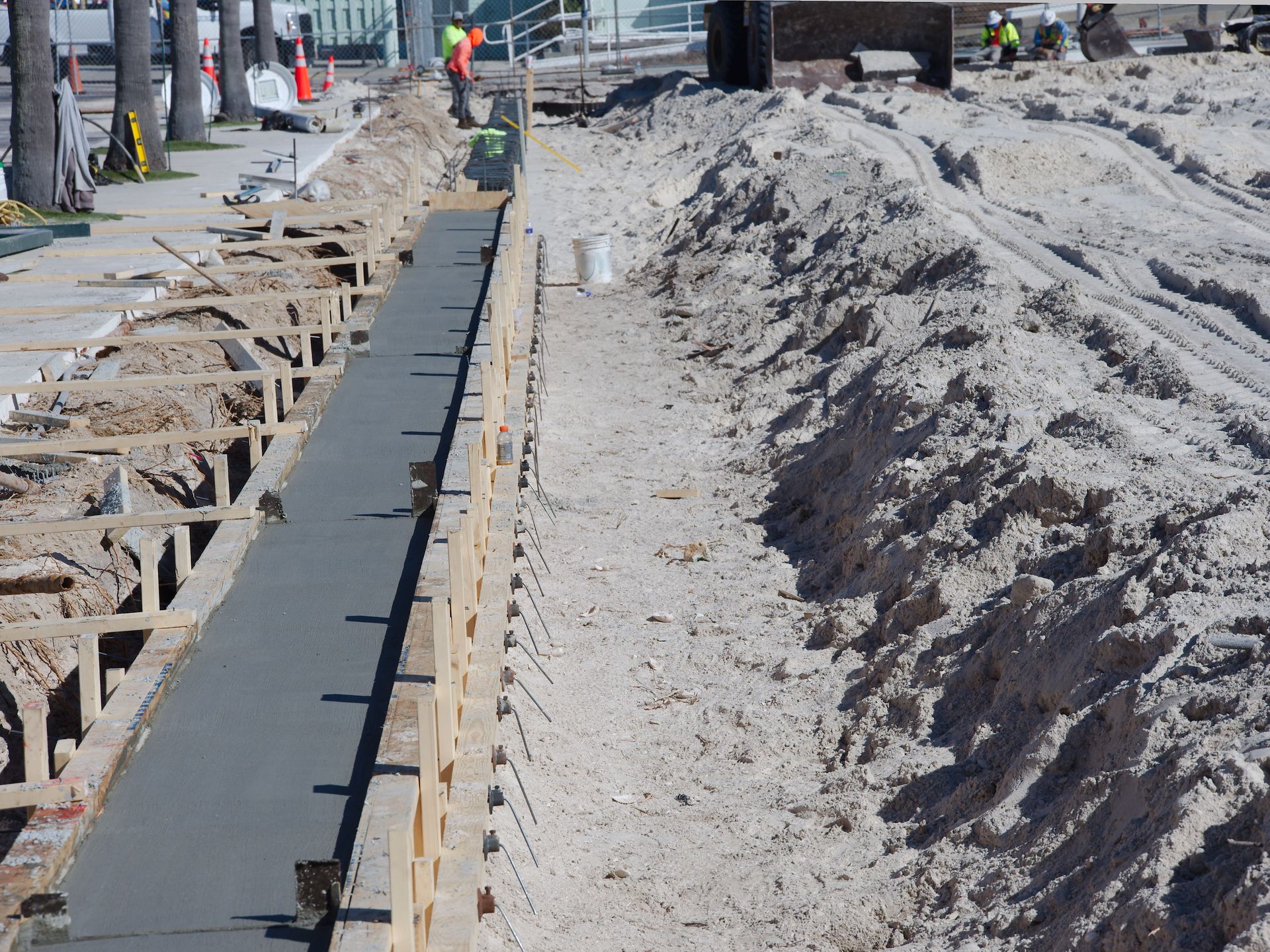 Road construction trench with fresh concrete curb beside a sidewalk and sand piles, workers in the distance