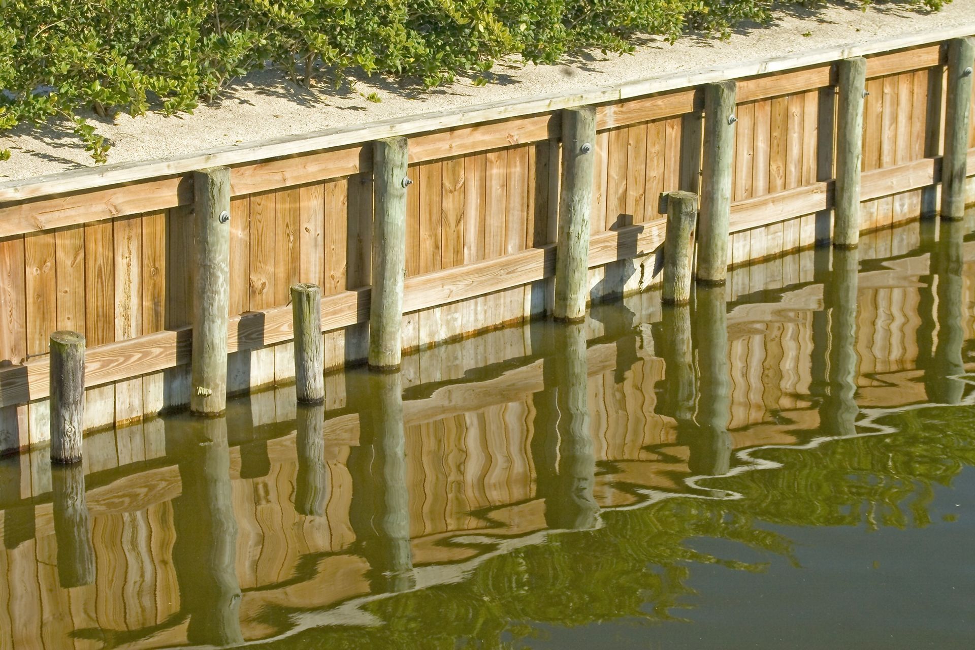 Wooden retaining wall along calm water, with reflections and a grassy bank above.