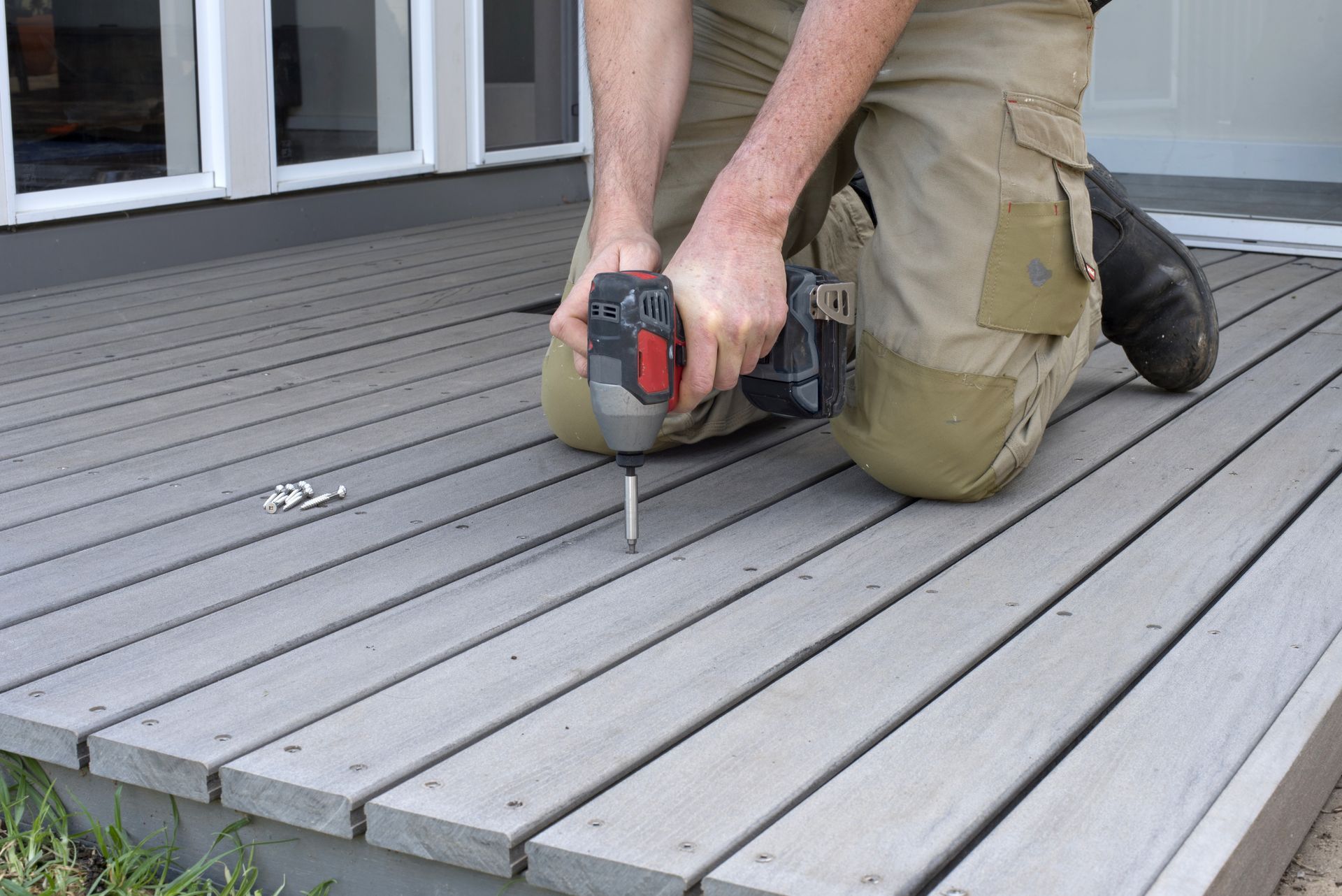 Person kneeling on a gray deck, using a drill to fasten a board near a house doorway
