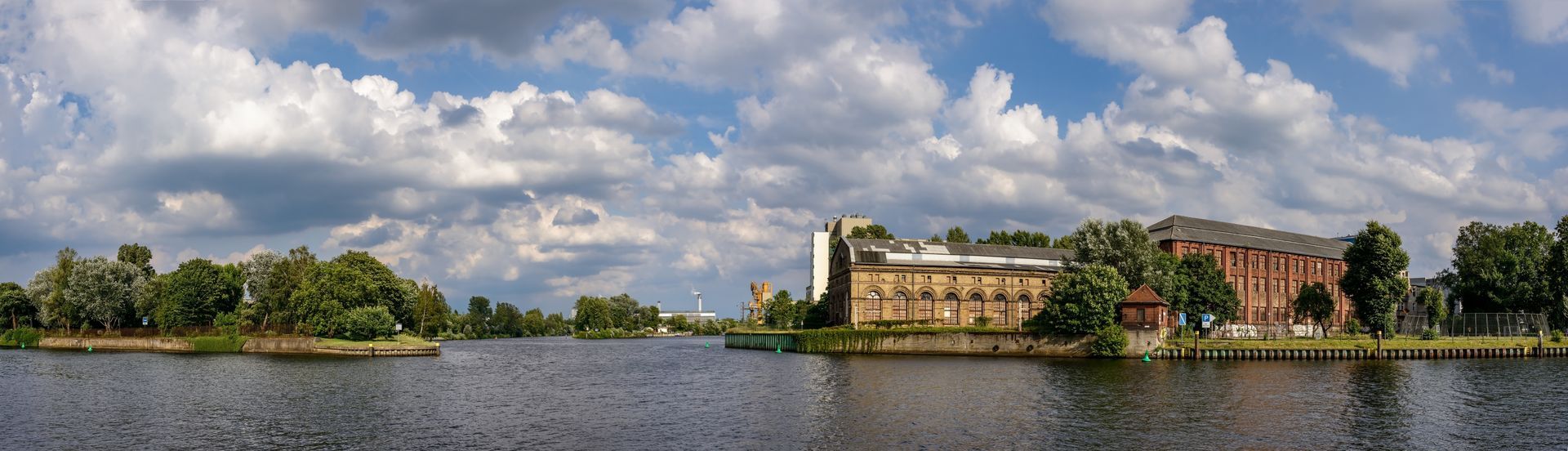 Wide river with a tree-lined shore and large buildings under a cloudy sky.