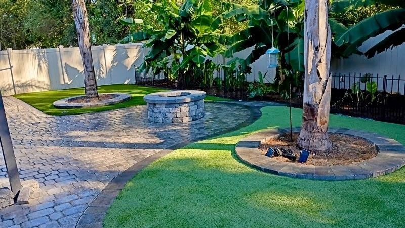 Backyard with fire pit, brick patio, artificial turf, and trees.