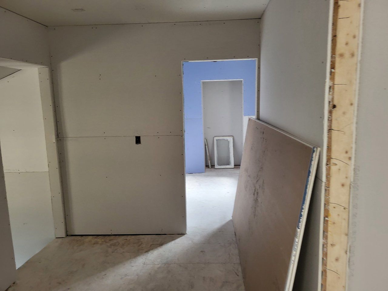 Unfinished interior room with drywall, leaning board, and doorway to a blue-walled adjacent space.