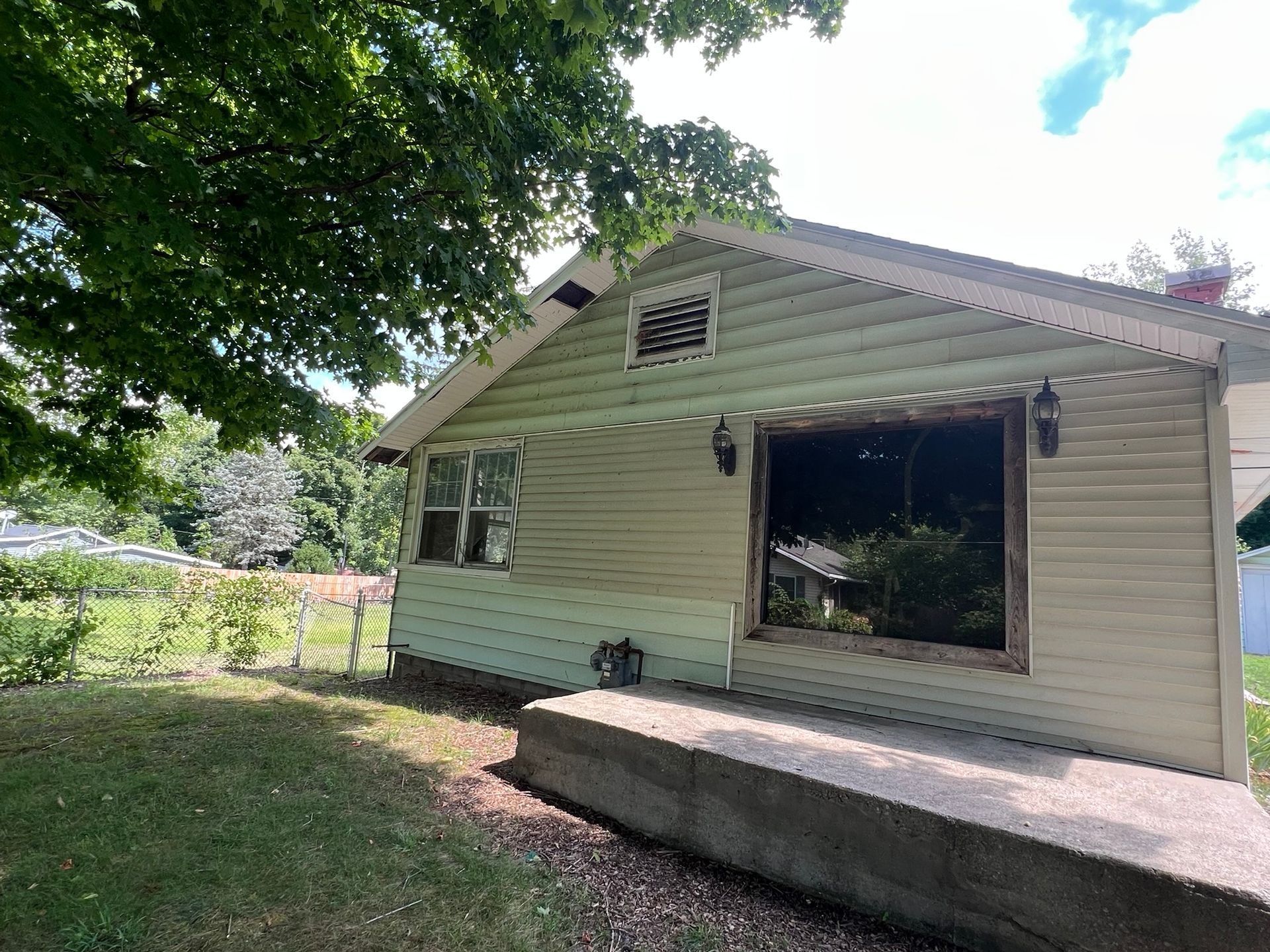 Small beige house with a broken front window and raised porch, viewed from a grassy yard under trees