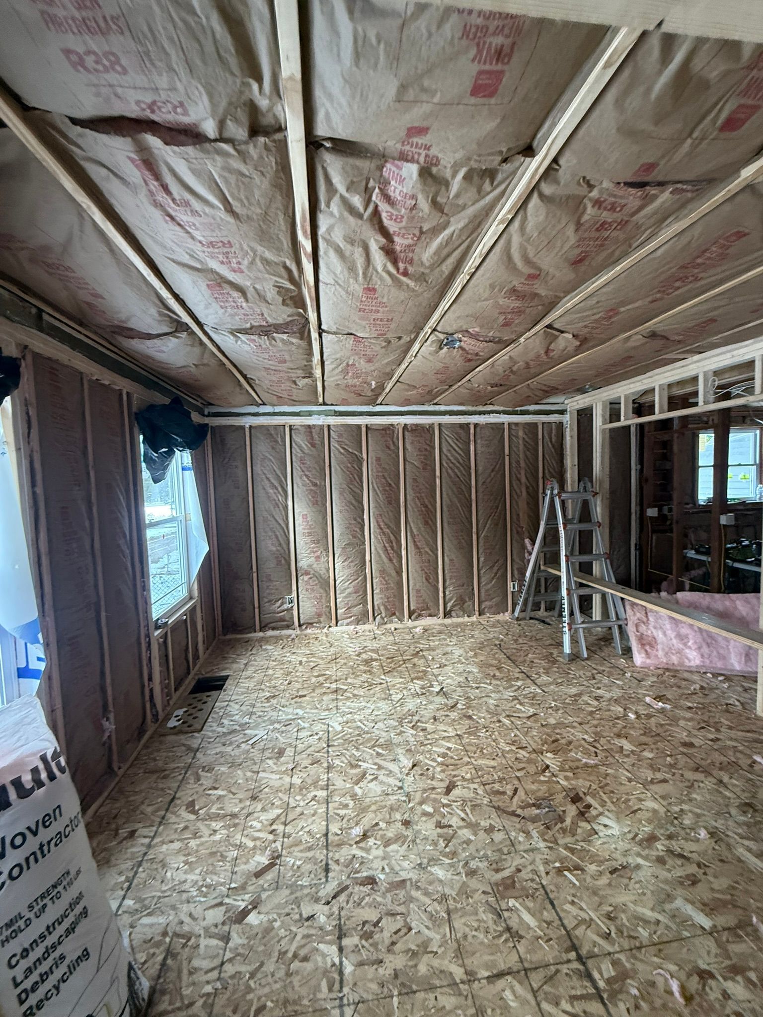 Unfinished room under construction with exposed insulation, wood framing, OSB floor, and a ladder near the back wall.