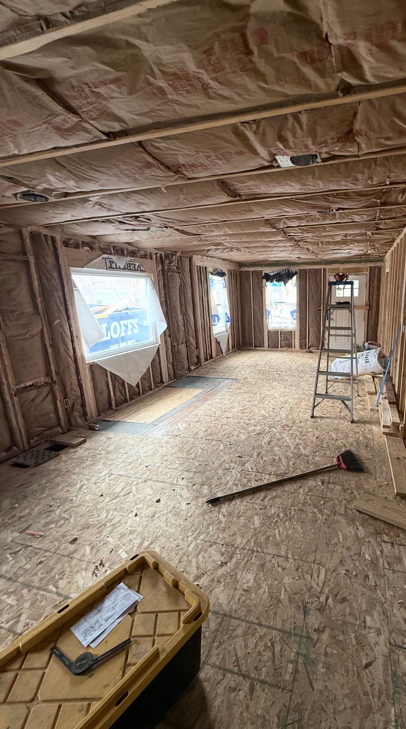 Unfinished room with exposed insulation, wood framing, debris on the floor, and a ladder by the wall.
