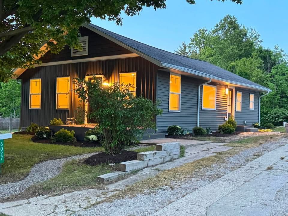 House with lit windows at dusk, gray siding, front porch, and a stone walkway in a quiet suburban setting