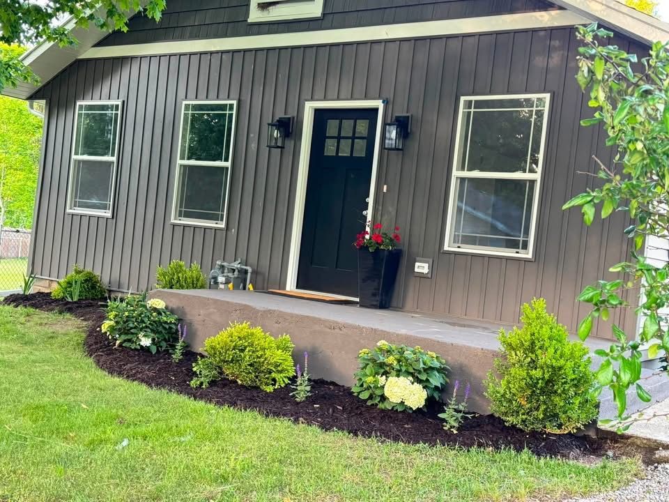 Small gray house with black door, front porch, and landscaped flower bed under green trees.