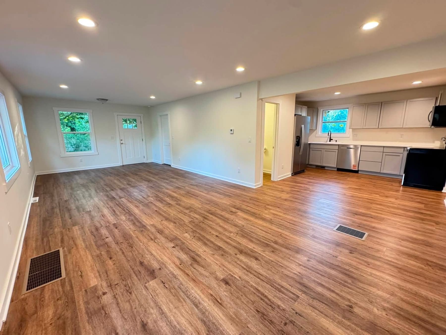 Empty living room with wood floors, recessed lights, and a small kitchenette area.