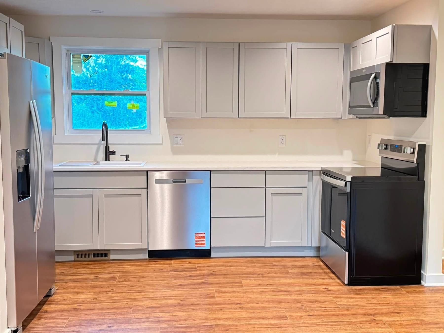 Small modern kitchen with gray cabinets, stainless appliances, white countertops, and a window above the sink.