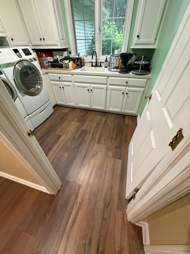 Small laundry room with white cabinets, washer, sink, and wood-look floor viewed through a doorway