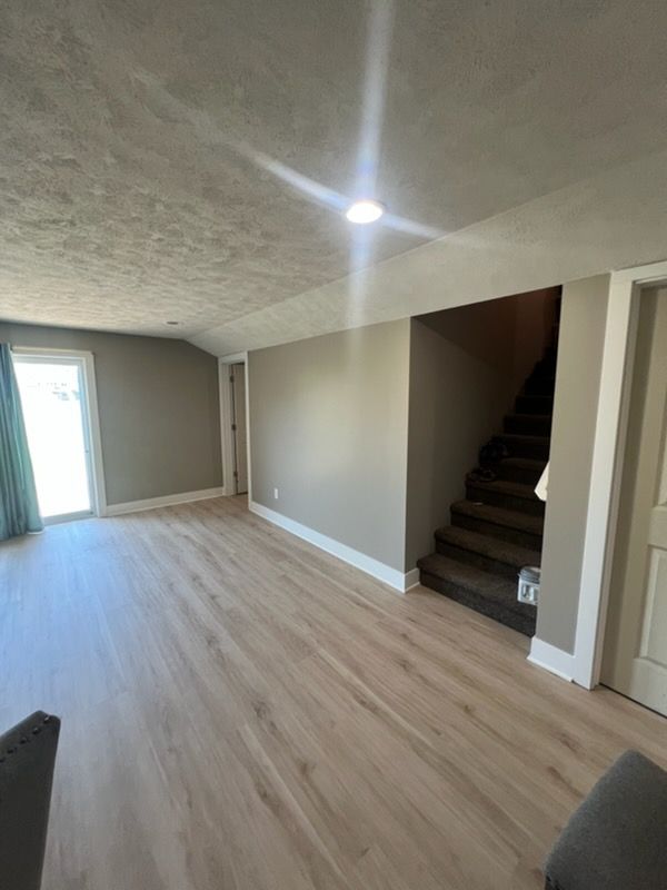 Empty living room with light wood floors, staircase, and bright doorway to the left