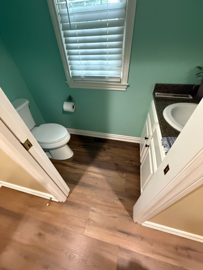 Small bathroom with green walls, wood floor, toilet, sink cabinet, and window with blinds.