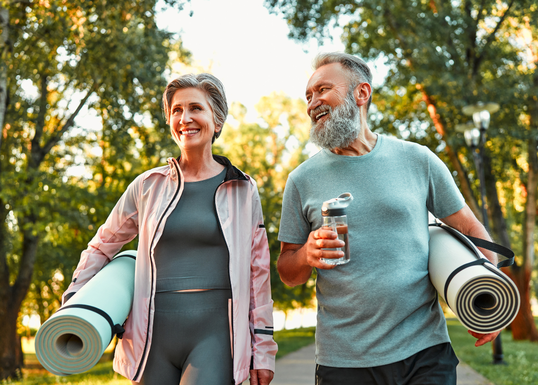 Older couple outdoors holding yoga mats, smiling.