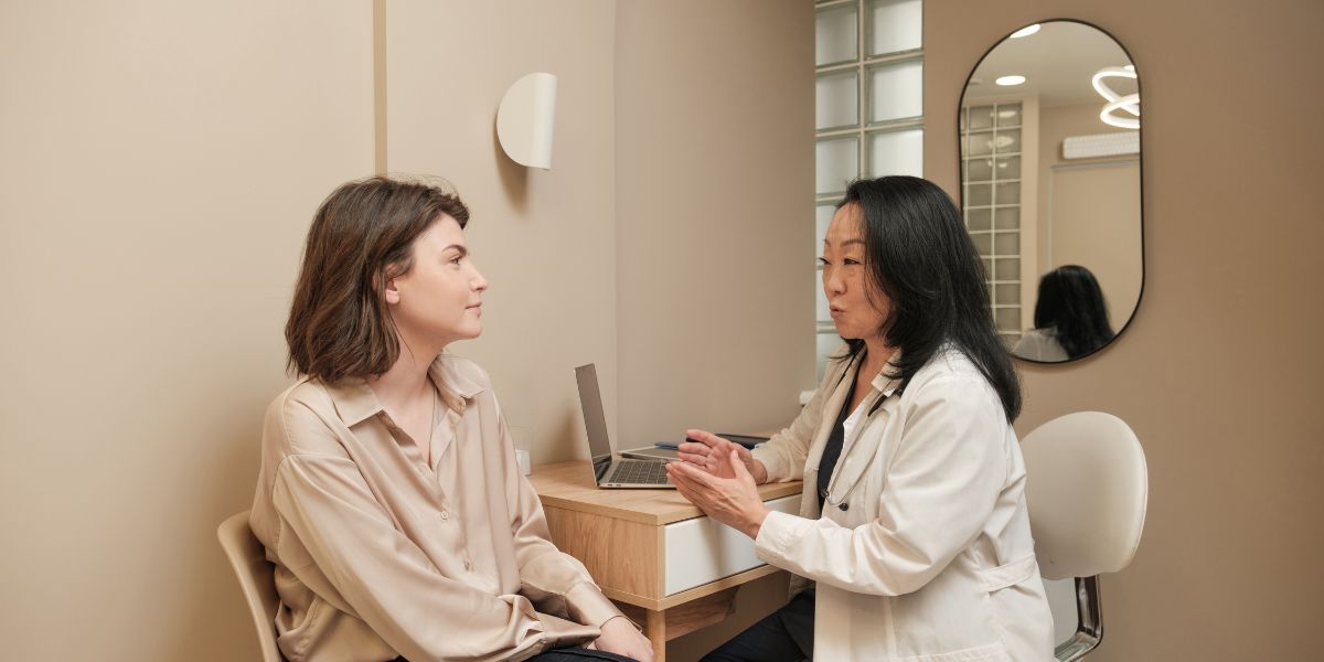 Two women talking in a bright room with a table, laptop, and mirror in the background