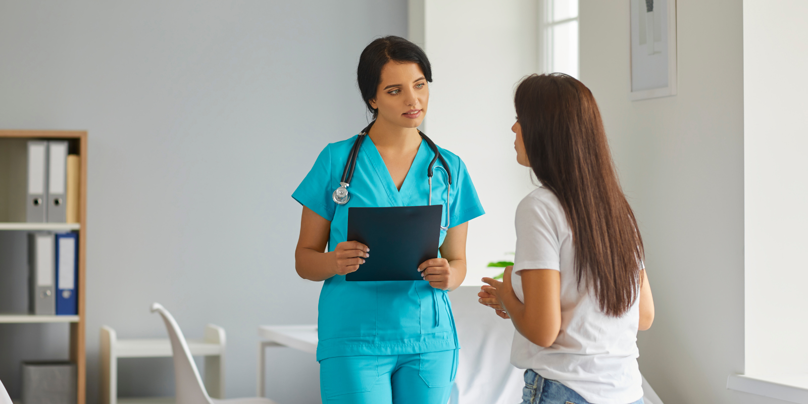 Healthcare worker speaking with a patient in a bright clinic office, holding a clipboard.